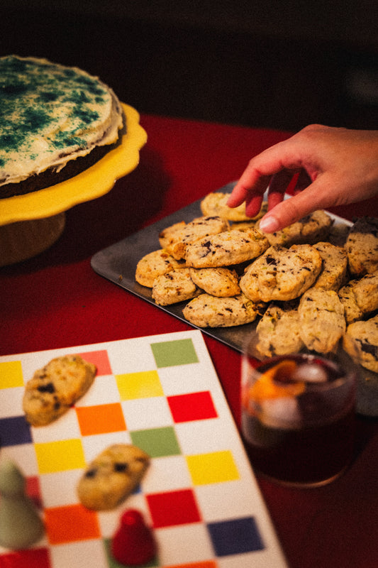 Hand reaching for cookies on a tray with a colorful chess board and a glass of negroni in the background