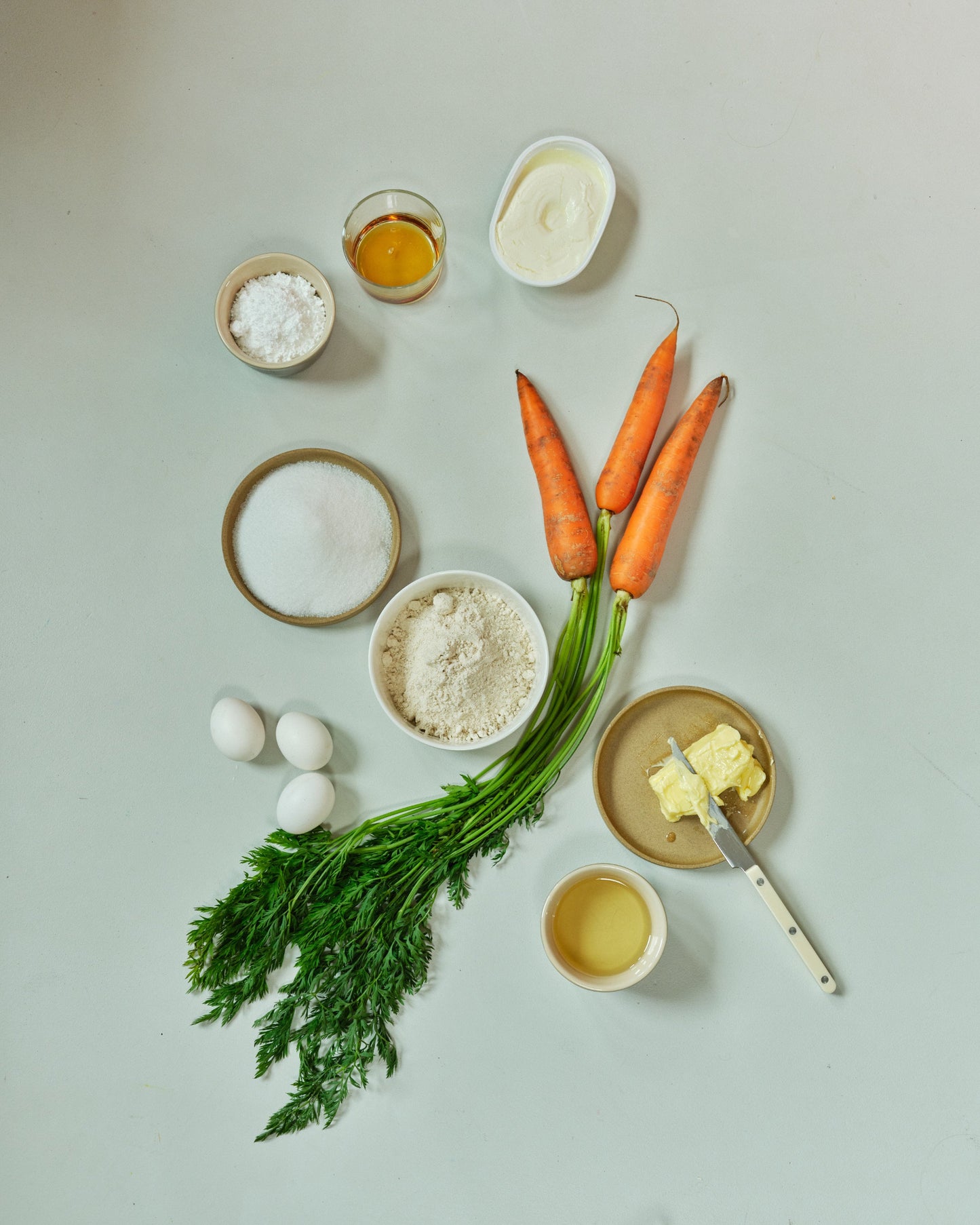 Ingredients for a carrot dish including carrots, eggs, flour, salt, and butter on a light background